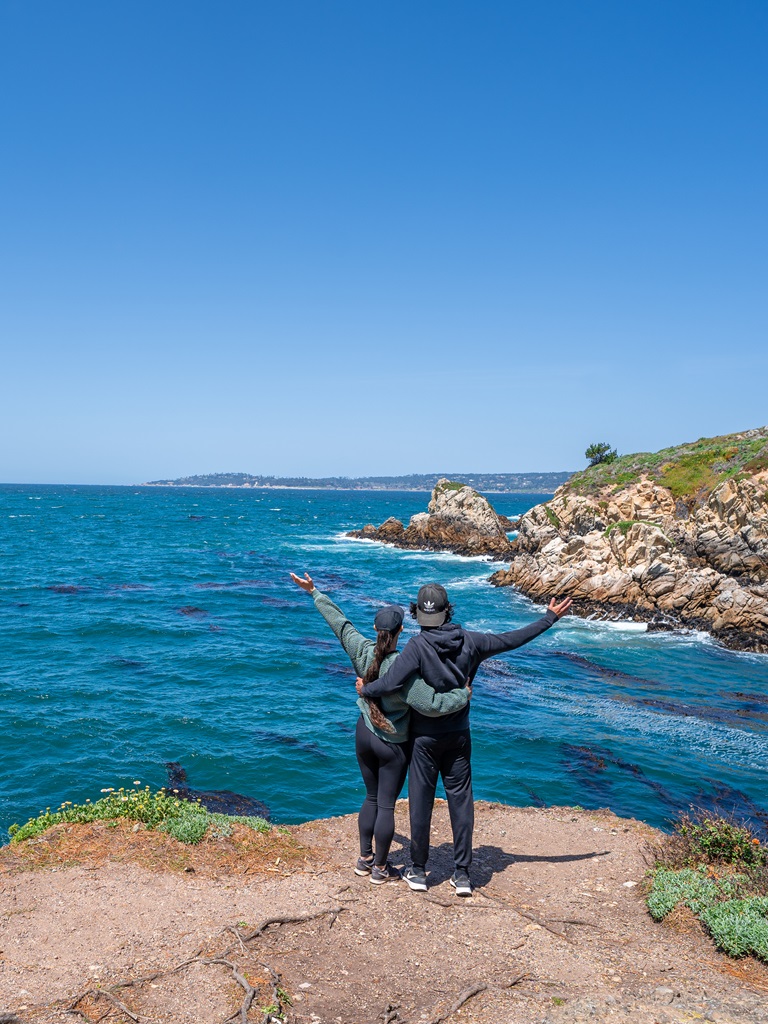 Man and woman standing with arms in the air at Coal Chute Point admiring the Pacific Ocean.