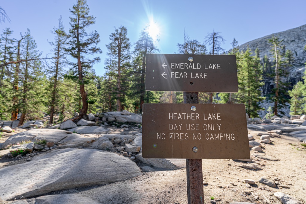 Two trail signs on one pole with top sign showing two arrows pointing to the left towards Emerald and Pear Lake, and bottom sign indicating Heather Lake - Day Use Only.