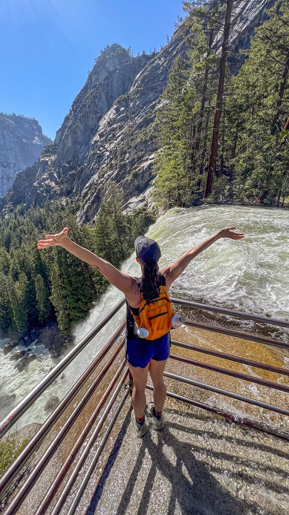 Woman standing at the top of Vernal Fall in Yosemite with hands in the air.