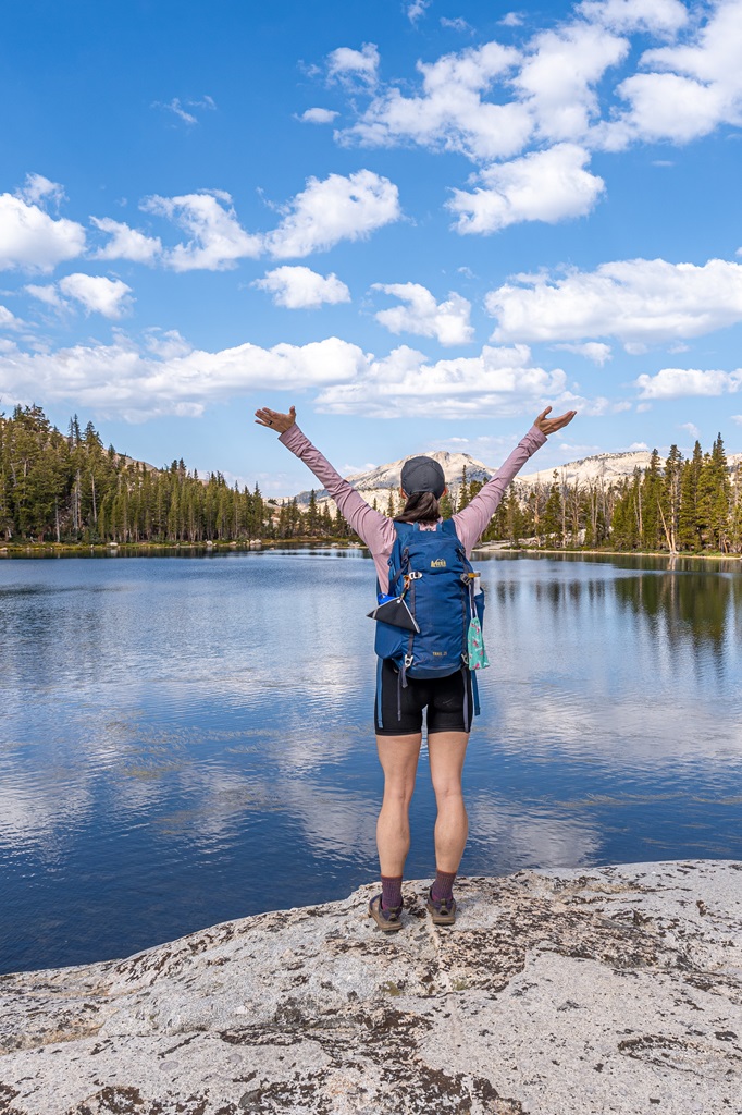 Woman standing on the granite shoreline of Lower Cathedral Lake with hands in the air.