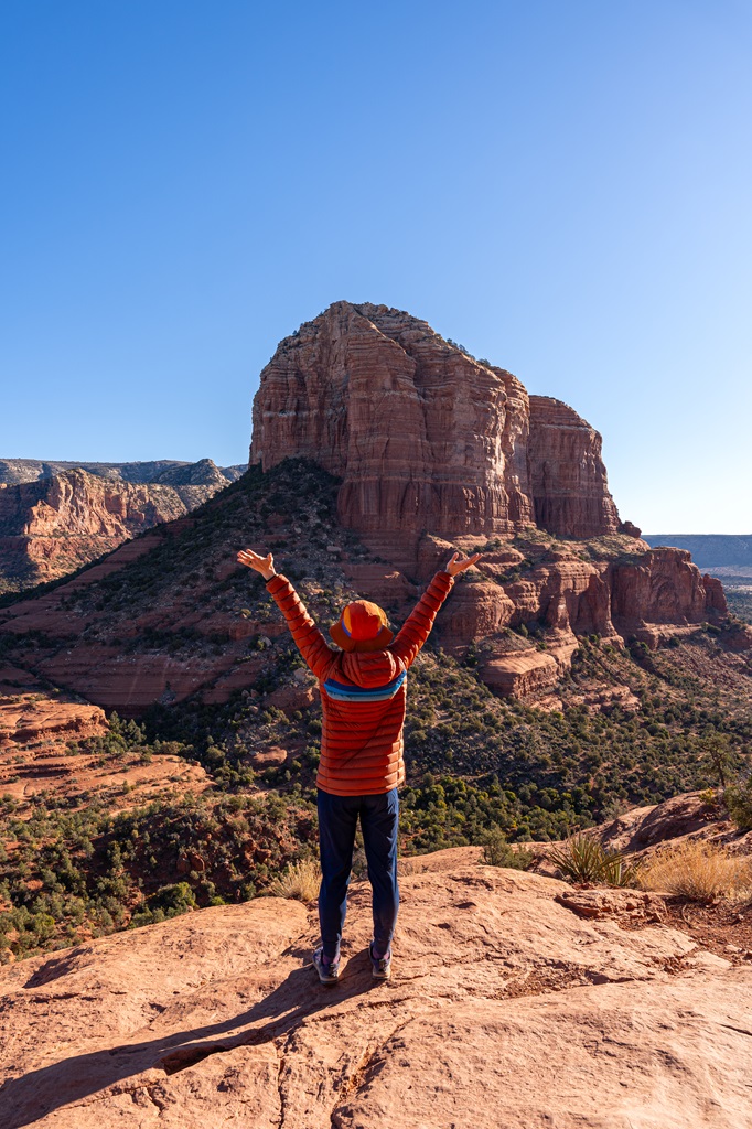 Woman with hands in the air standing on the upper section of the Bell Rock Climb in Sedona with red rock formations in the distance.