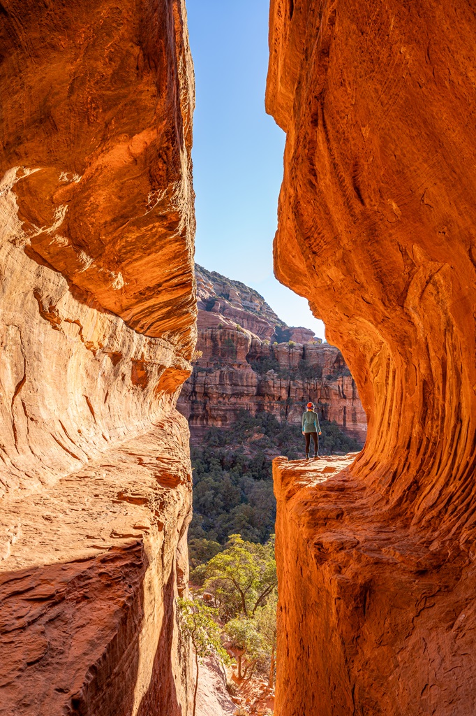 Woman standing on the right side inside the Subway Cave with the sun shining making the cave glow a bright orange.