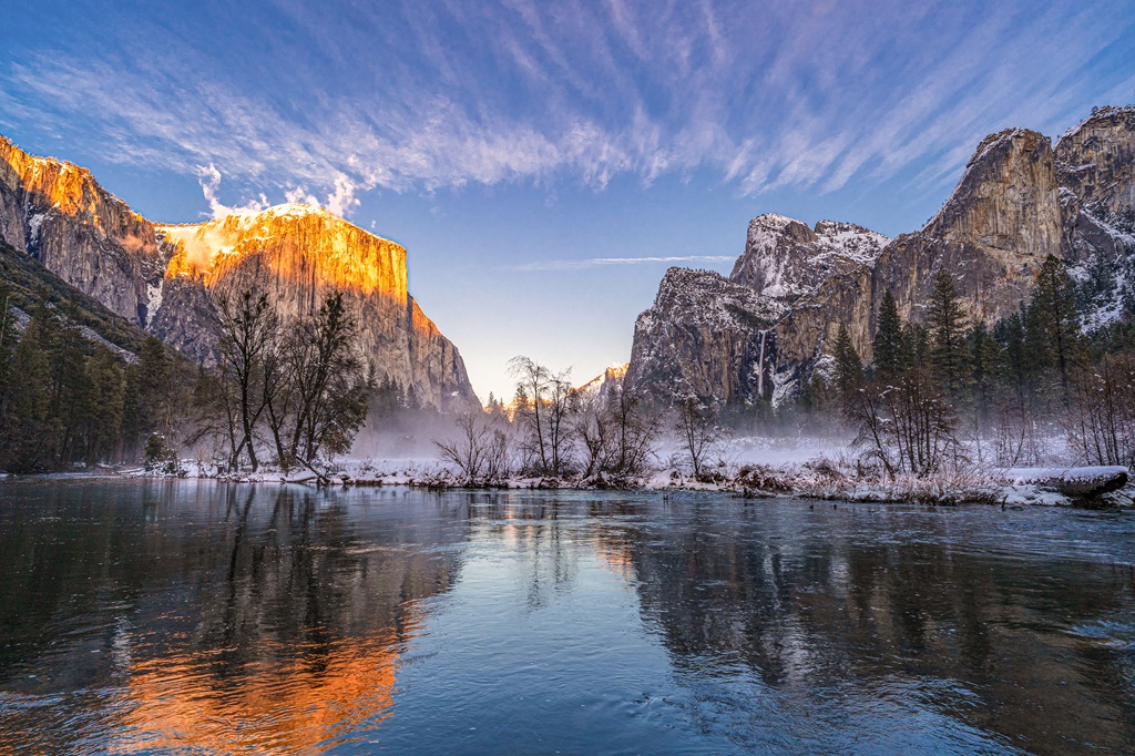 Sun setting at Valley View in Yosemite National Park during winter with a light fog hovering over the ground and reflections of El Capitan and other snowy granite peaks.