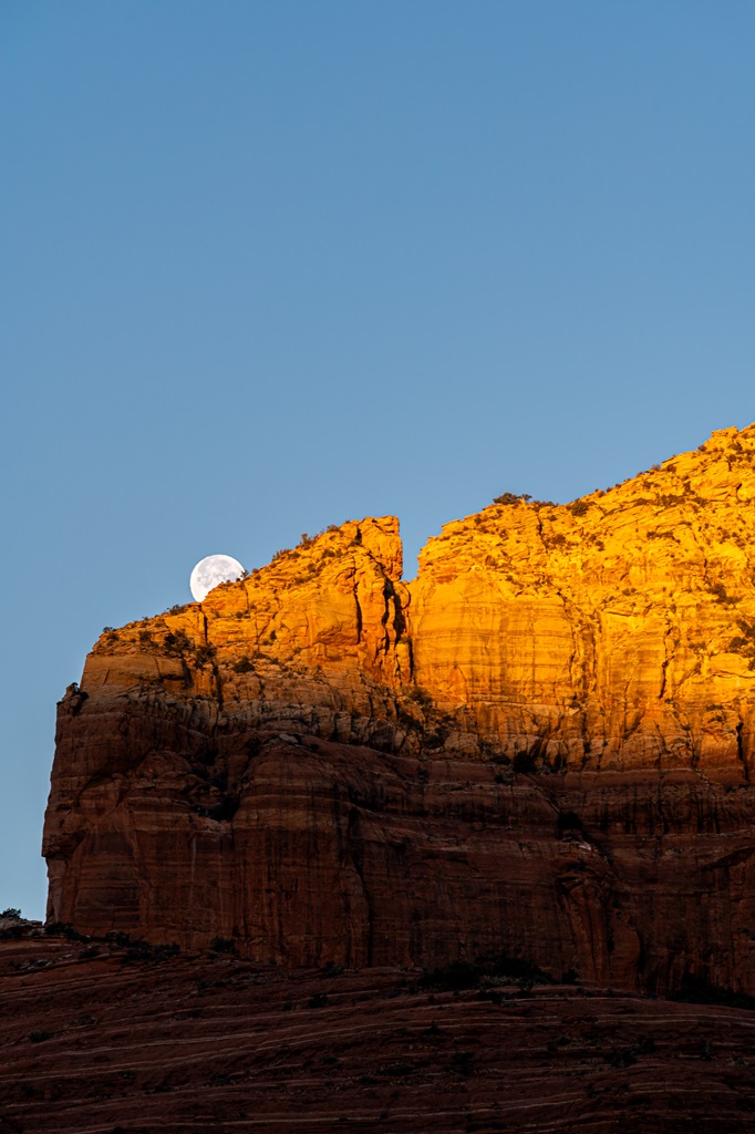 Full moon halfway behind a red rock formation with the sunrise light shining on the rocks.