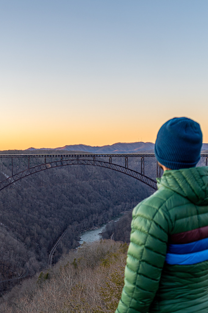 Man standing at Long Point Overlook watching the sunset with the horizon a faint orange color and view of the New River Gorge Bridge. 