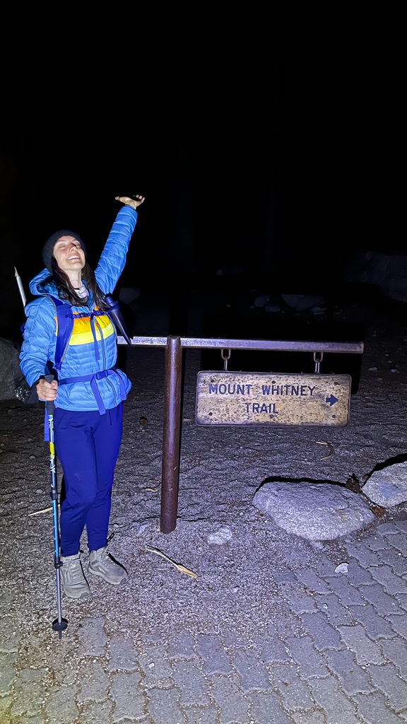 Woman posing beside the Mount Whitney Trailhead sign at Whitney Portal at nighttime.