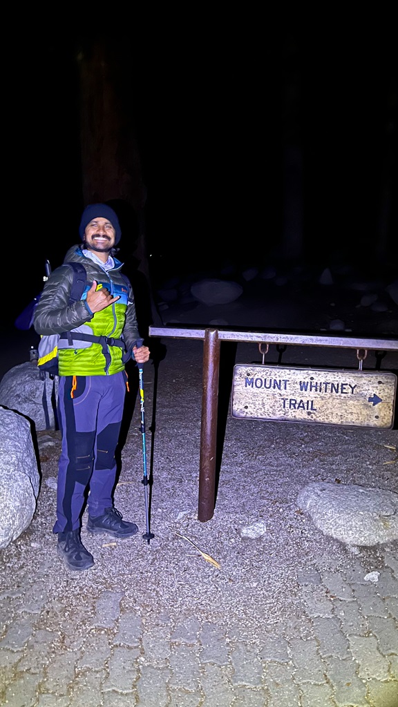 Man posing beside the Mount Whitney Trailhead sign at Whitney Portal at nighttime.