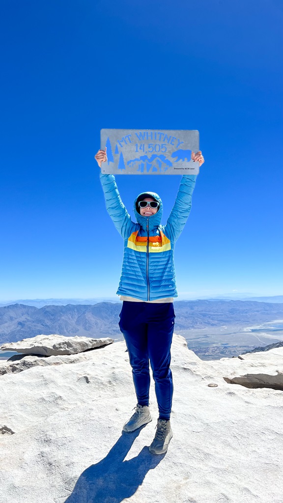 Woman holding the Mt Whitney summit sign on top of Mt Whitney.