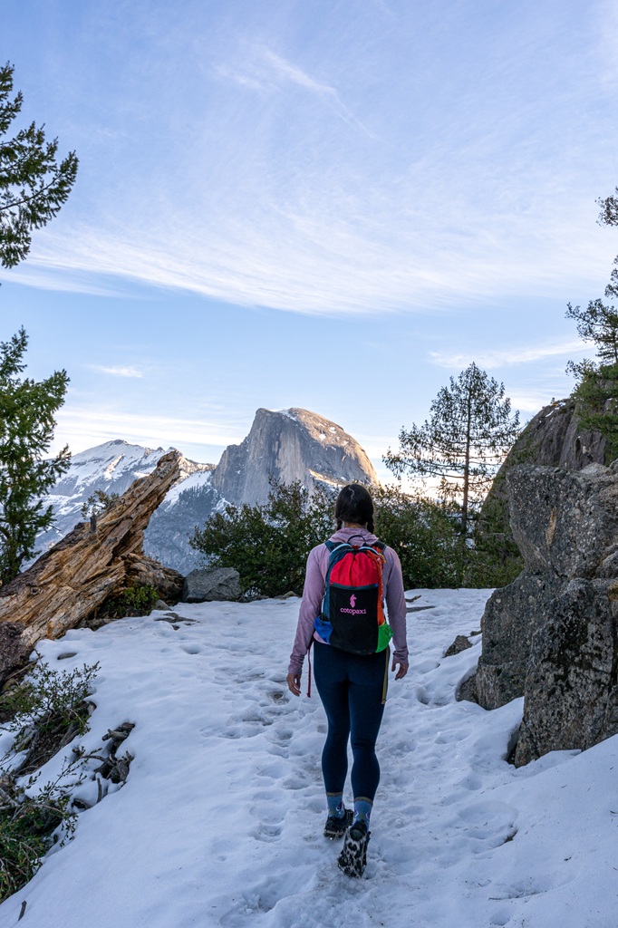 Woman hiking along a snow-covered section on the Four Mile Trail with Half Dome in the background.