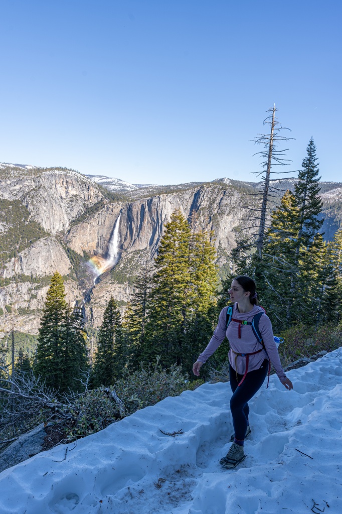Woman hiking along the Four Mile Trail in snow in Yosemite National Park with Yosemite Falls in the background.