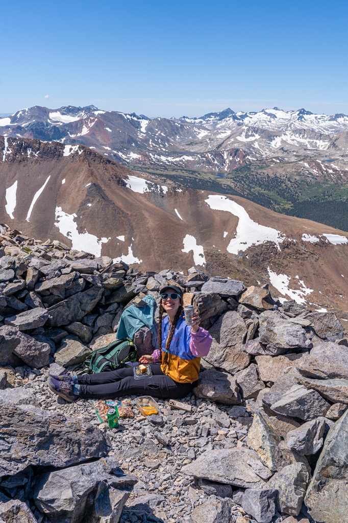 Woman sitting behind a small rock wall, eating snacks and drinking an Olipop on the summit of Mount Dana.