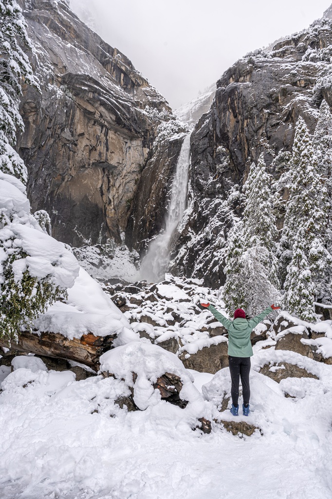 Woman standing on a rock looking at Lower Yosemite Fall from a snow-covered viewpoint in mid-winter.