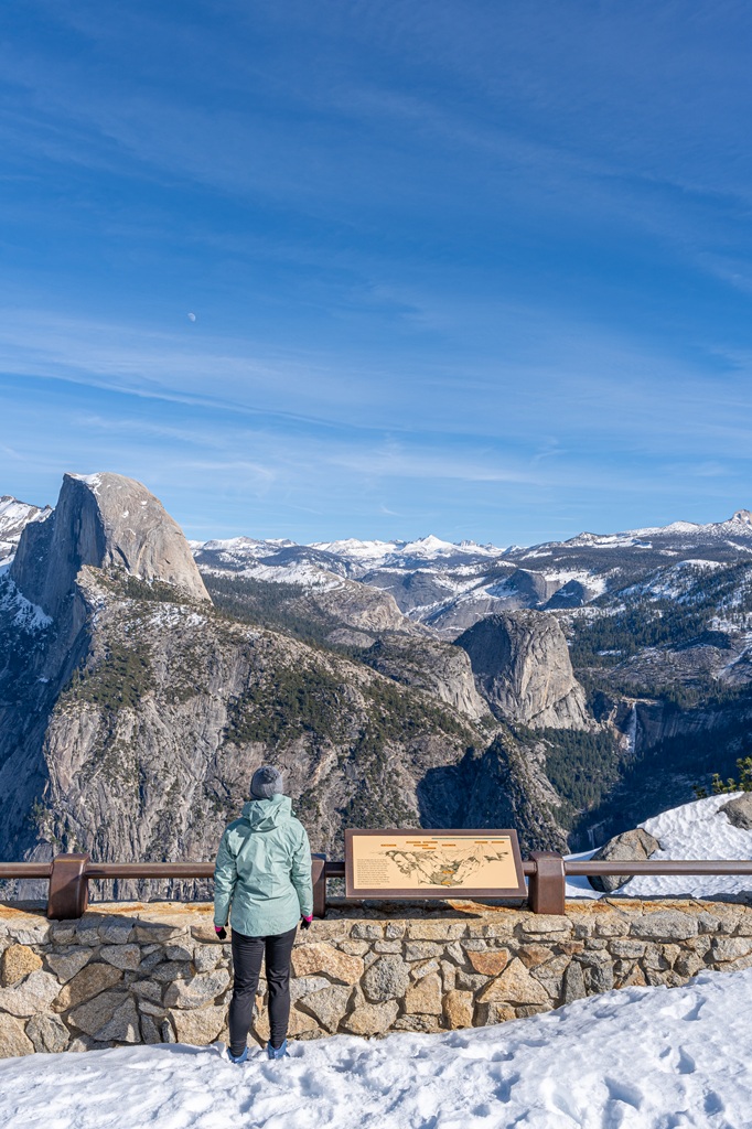 Woman standing at Glacier Point Overlook in snow during winter looking at Half Dome and snow-covered peaks in the distance.