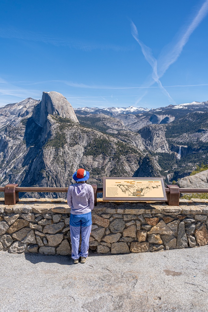 Woman standing at the Glacier Point Overlook looking at Half Dome, Nevada Fall, and snowy peaks in the distance in Yosemite National Park.