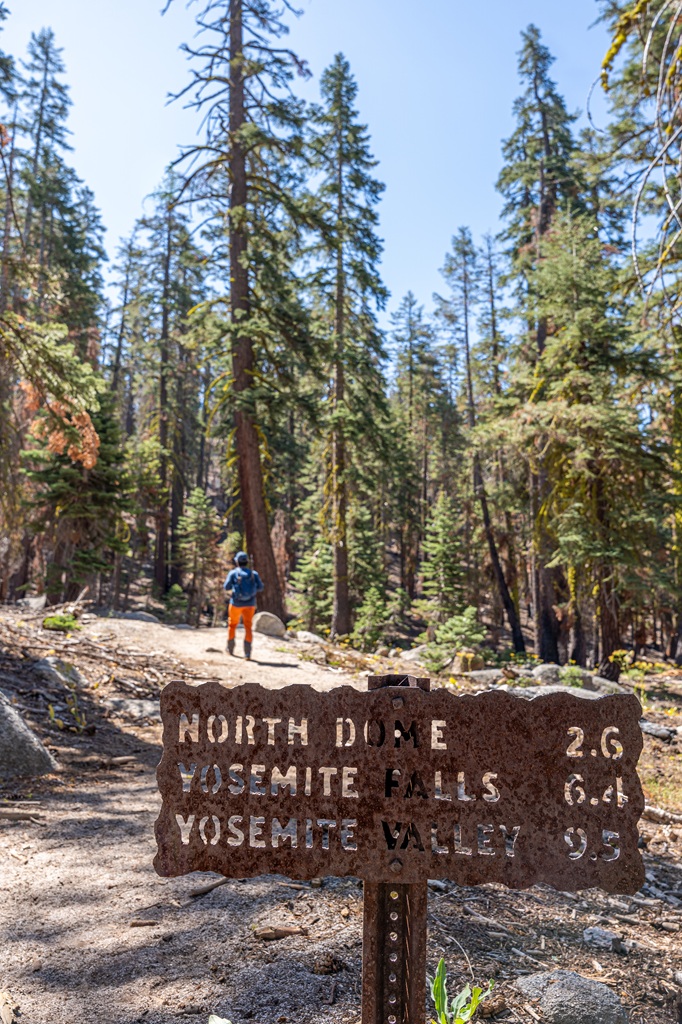 Trail sign indicating distance to North Dome, Yosemite Falls, and Yosemite Valley with a man hiking along the North Dome Trail in the background.