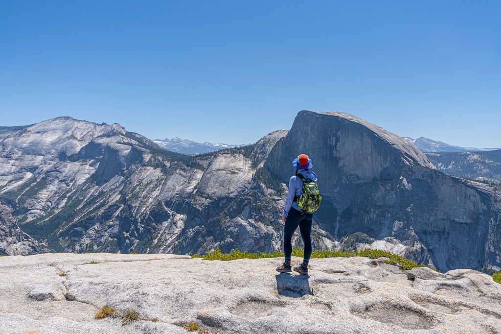 Woman standing on North Dome looking at the view of Half Dome and Clouds Rest in Yosemite National Park.