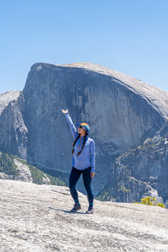 Woman posing for a picture from the top of North Dome with Half Dome in the distance.